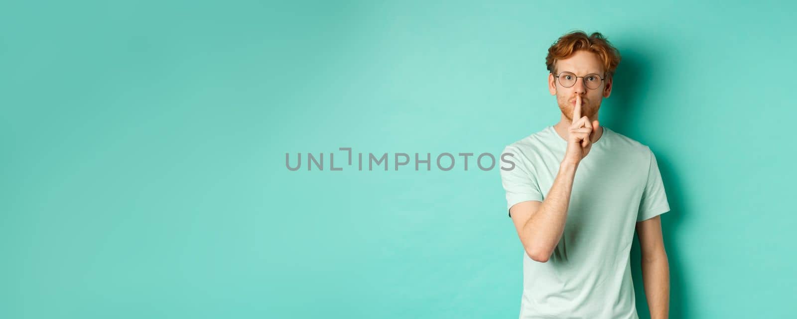 Serious redhead man hushing, tell to be quiet, making shush gesture and looking at camera, standing in t-shirt over turquoise background.