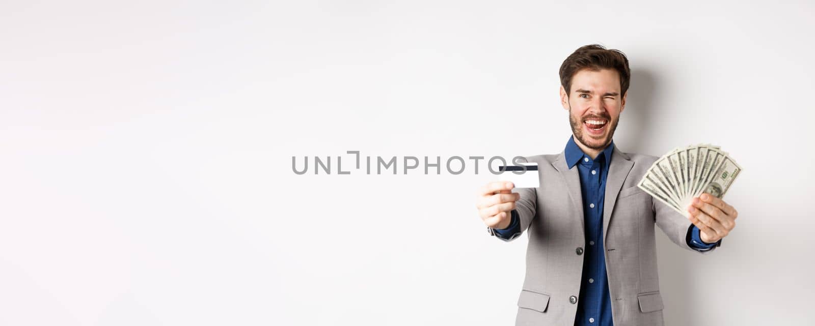 Happy successful businessman in suit showing money and credit card, smiling at camera, white background.