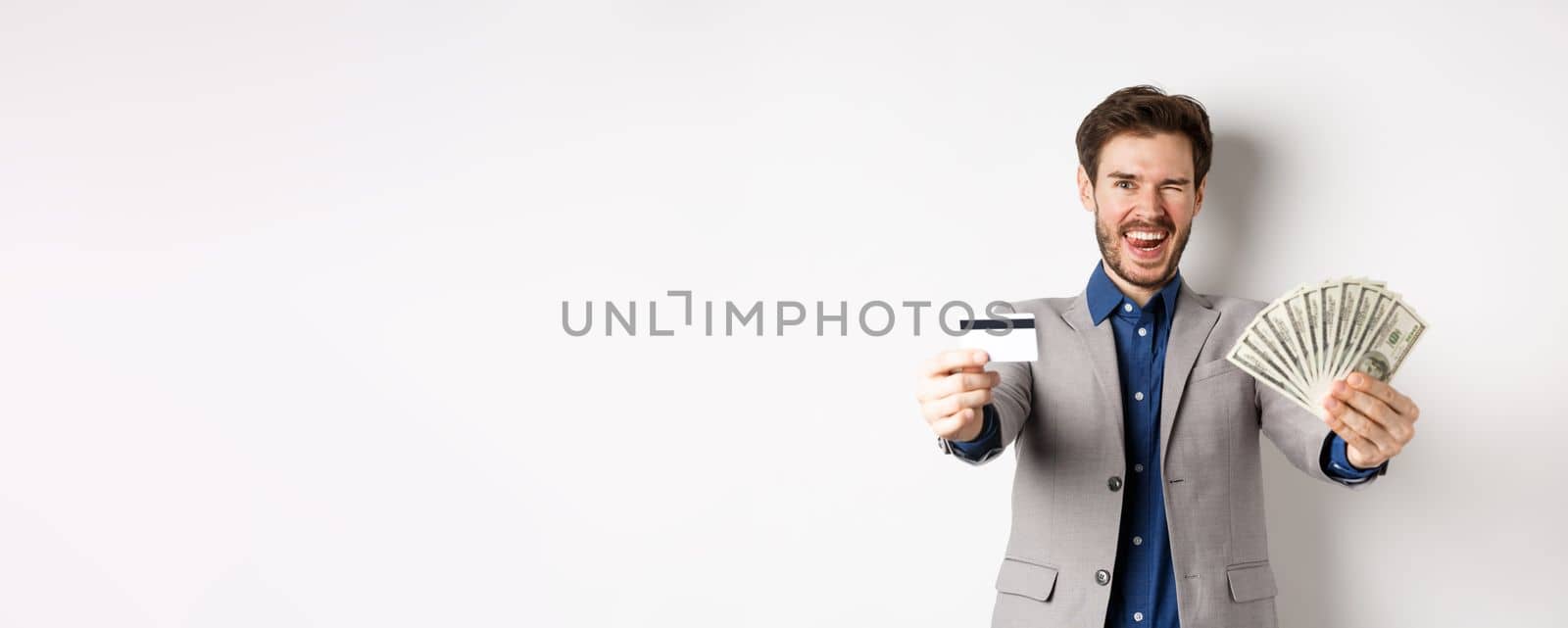 Happy successful businessman in suit showing money and credit card, smiling at camera, white background by Benzoix