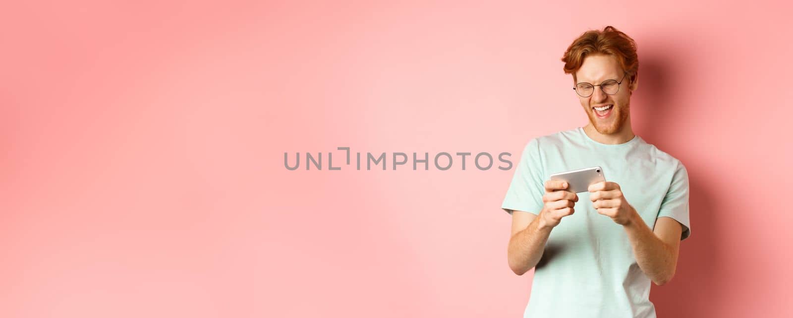 Happy young man with red messy haircut, wearing glasses, playing video game on smartphone and having fun, looking at mobile screen, standing over pink background by Benzoix