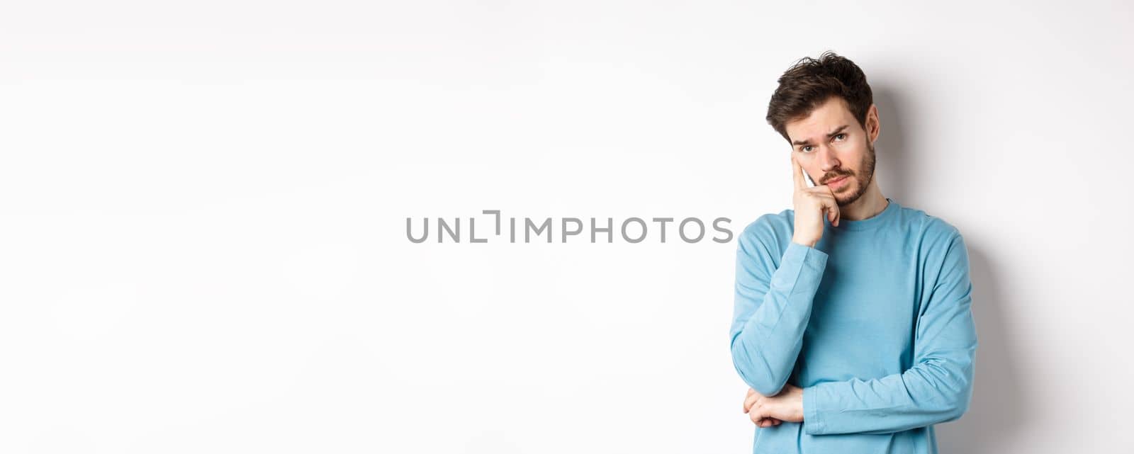 Skeptical young man listening to you with bored face, looking at camera reluctant, standing in sweatshirt over white background.