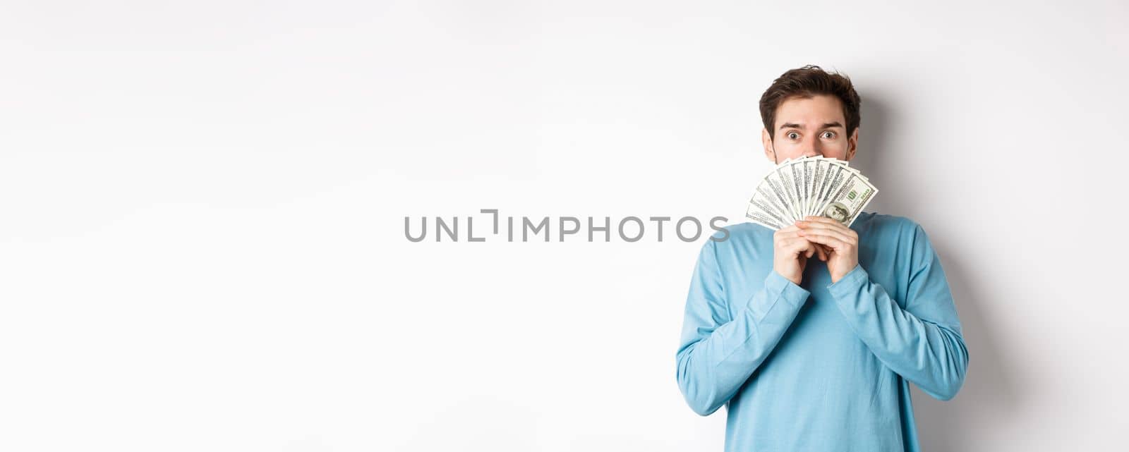 Surprised and amazed handsome guy showing money, looking at promo offer, going shopping with cash, standing over white background.