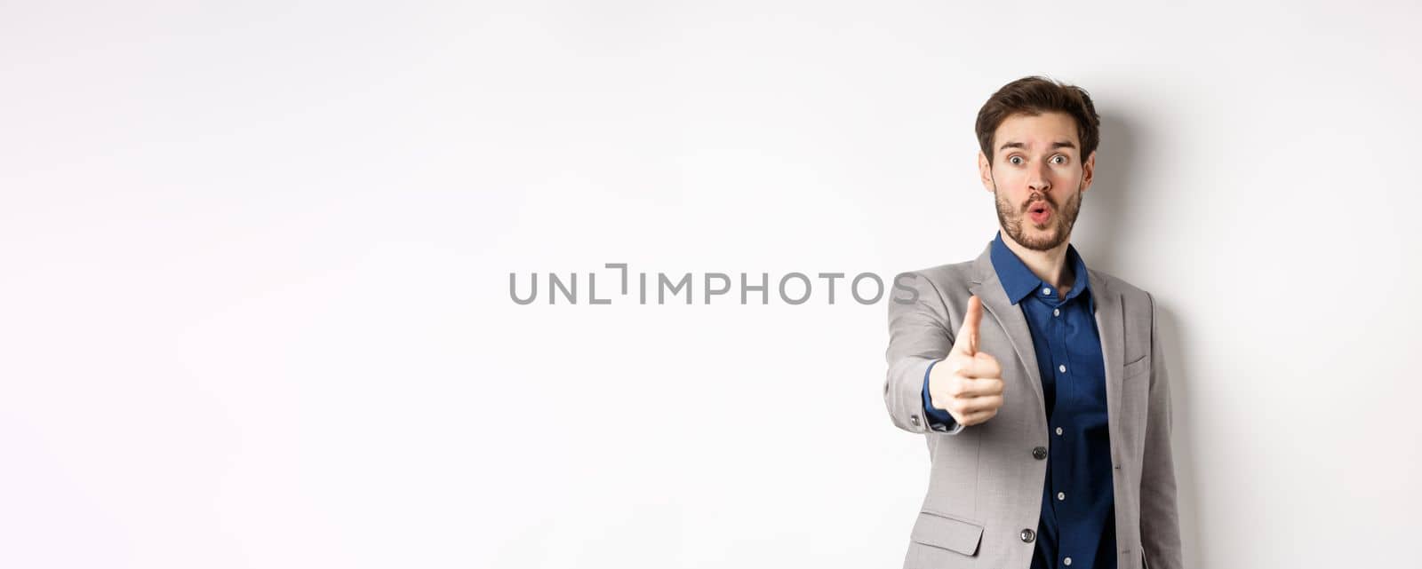 Image of handsome businessman in suit showing thumb up with excited face, standing on white background.