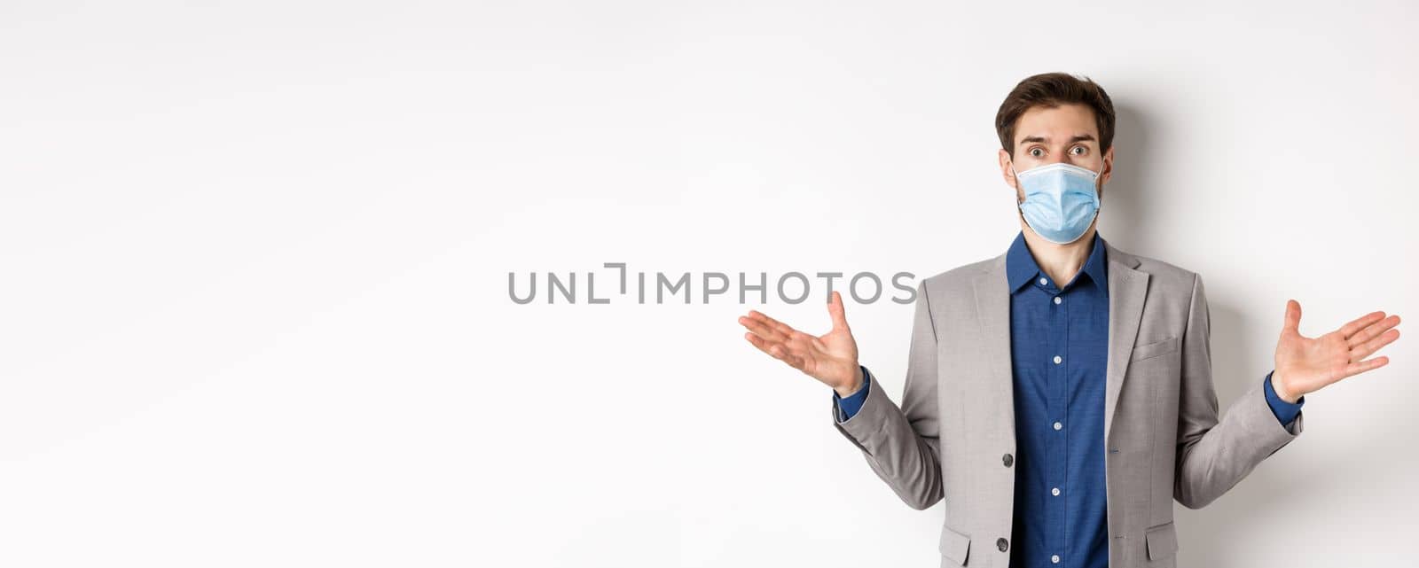 Health, pandemic and business concept. Confused businessman in medical mask and suit spread hands sideways and look puzzled, cant understand, white background.