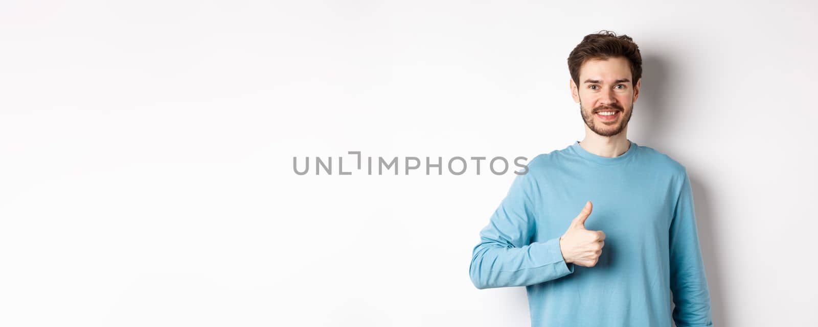 Smiling male model in casual shirt showing thumb up, approve and recommend product, praise excellent choice, standing on white background.