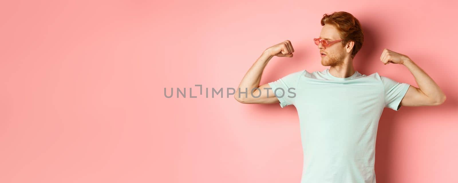 Confident young man with red hair, wearing summer sunglasses and t-shirt, showing strong and fit body muscles, flex biceps and staring cool at camera, pink background. Workout and gym concept