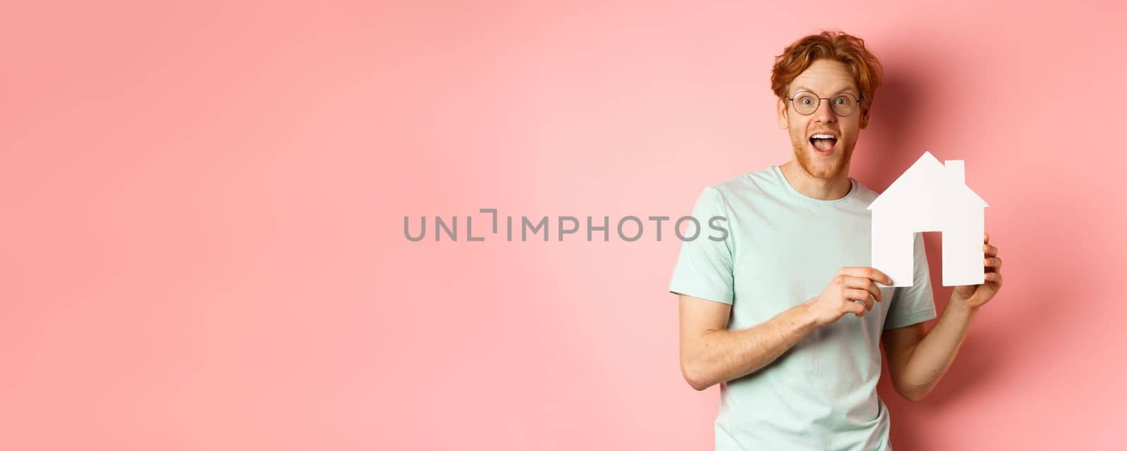 Real estate. Cheerful redhead man buying property, showing paper house cutout and smiling amazed, standing over pink background by Benzoix