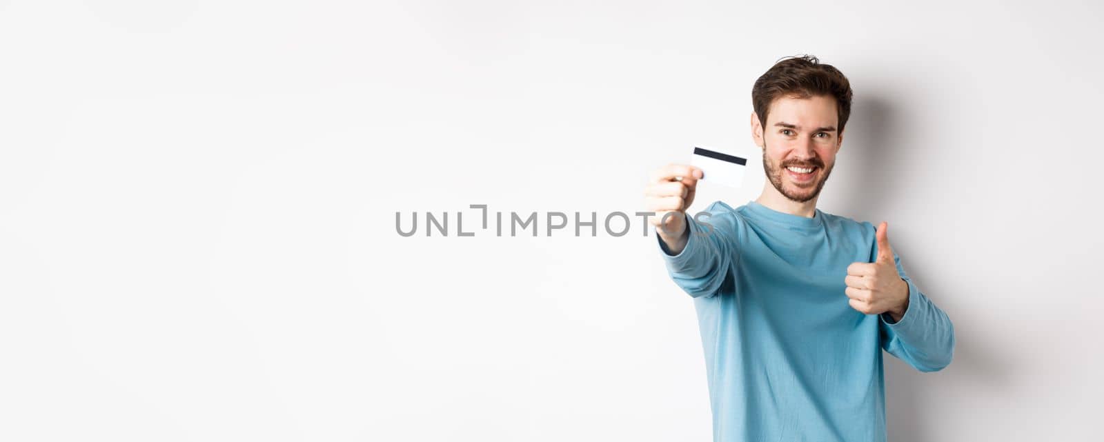 Handsome caucasian man showing plastic credit card with thumb-up, recommend and praise good bank offer, standing on white background. by Benzoix