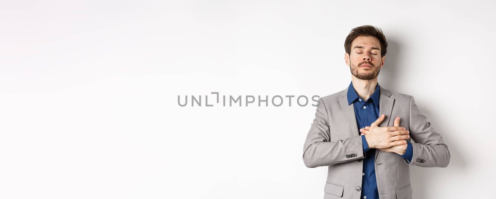 Young man in suit close eyes, holding hands on heart and remember happy memories, feeling nostalgic, standing against white background by Benzoix