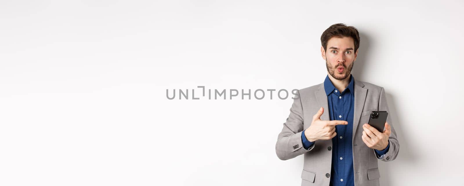 Excited guy in suit say wow, found something online on smartphone, pointing at mobile phone impressed, white background by Benzoix