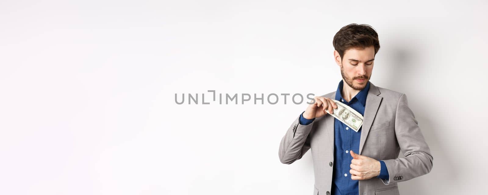 Successful businessman put dollars inside suit pocket, hiding money, standing on white background.