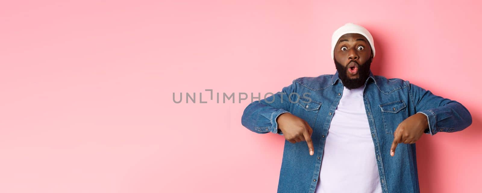 Amazed and impressed african-american guy showing advertisement, staring at camera happy and pointing fingers down, pink background.