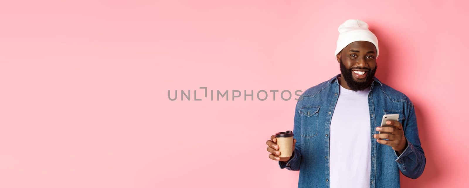 Image of stylish Black man hipster drinking takeaway coffee, reading message on phone and smiling, standing over pink background.