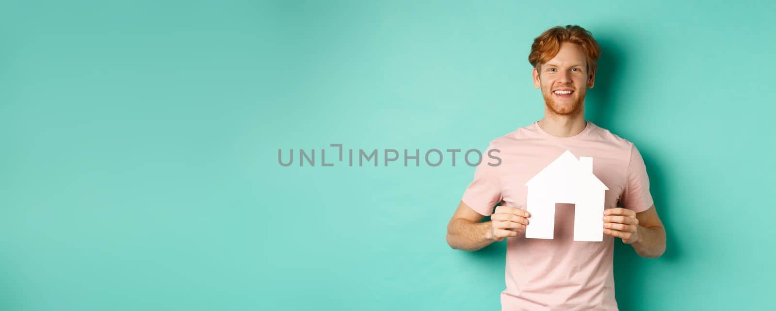 Real estate concept. Young man with red hair, wearing t-shirt, showing paper house cutout and smiling happy, standing over mint background. Copy space