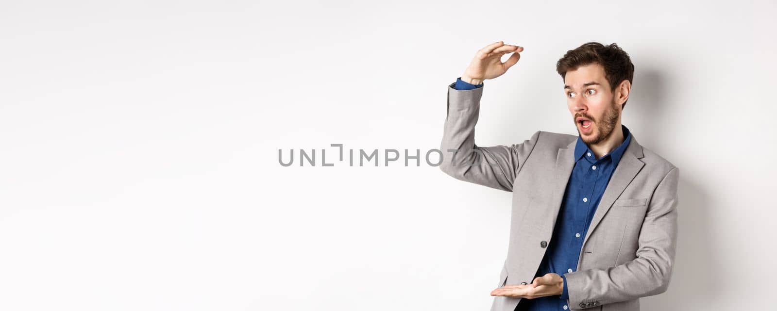 Handsome salesman in suit showing big size, large profit, smiling at camera enthusiastic, standing on white background.