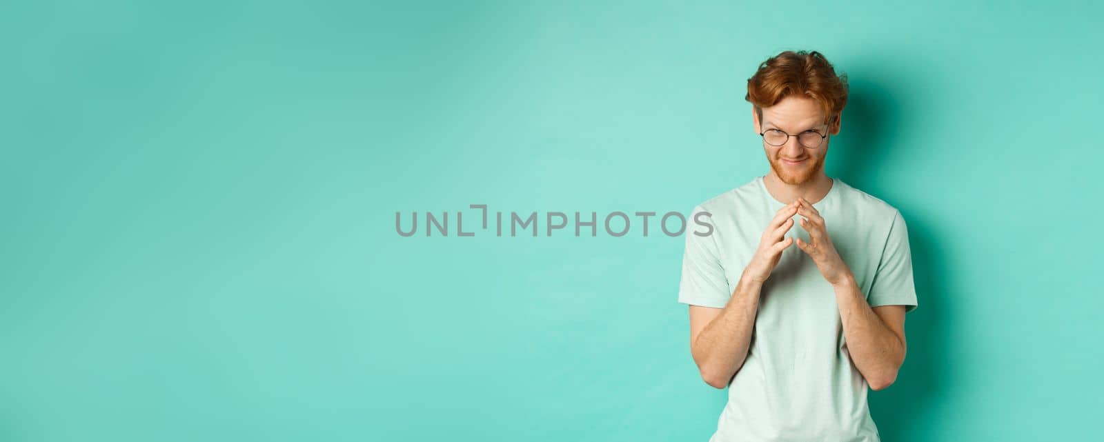 Devious redhead man in glasses and t-shirt pitching an idea, steeple fingers and look from under forehead with sly and smug smile, standing over mint background.
