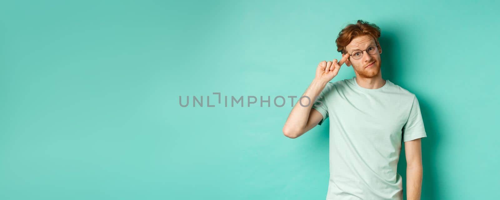 Skeptical redhead man scolding someone stupid or crazy, pointing finger at head and staring at camera, standing over mint background.