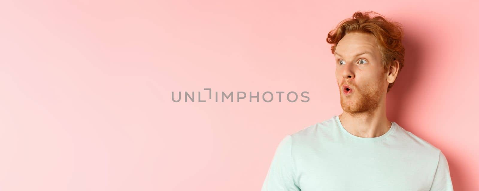 Headshot portrait of surprised redhead man with beard, looking left and saying wow, raising eyebrows amazed, standing over pink background.