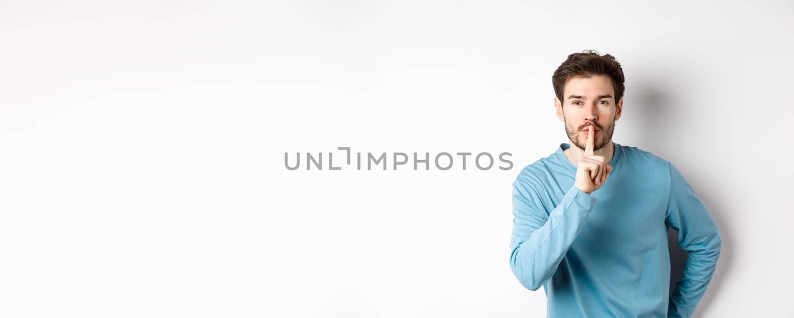 Attractive bearded man asking to keep quiet, showing taboo hush gesture and looking at camera calm, standing over white background by Benzoix