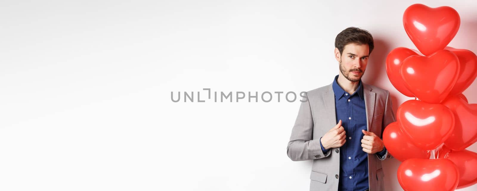 Lovers day. Handsome and confident young man getting dressed for Valentines day, fixing suit and looking at camera, standing near romantic heart balloons, white background.