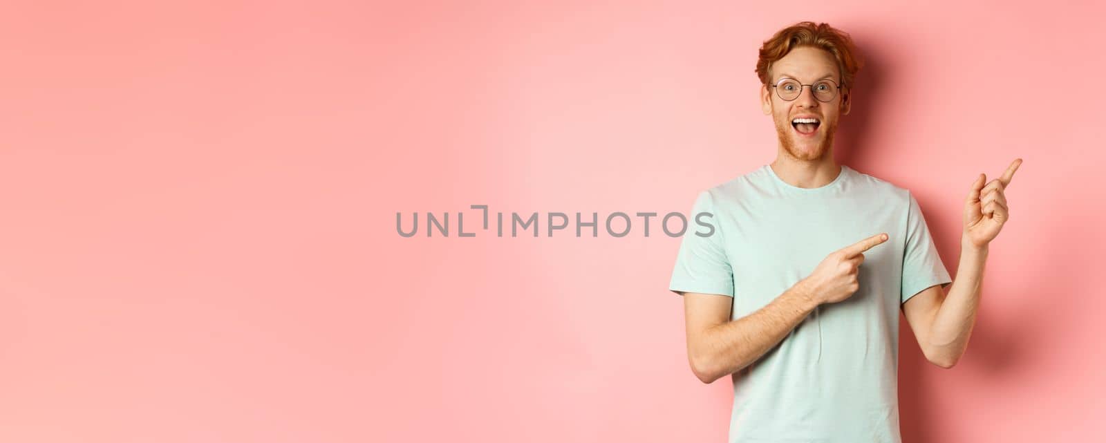 Amazed redhead man in glasses and t-shirt showing special promotion, pointing fingers at upper right corner and staring excited at camera, standing over pink background by Benzoix