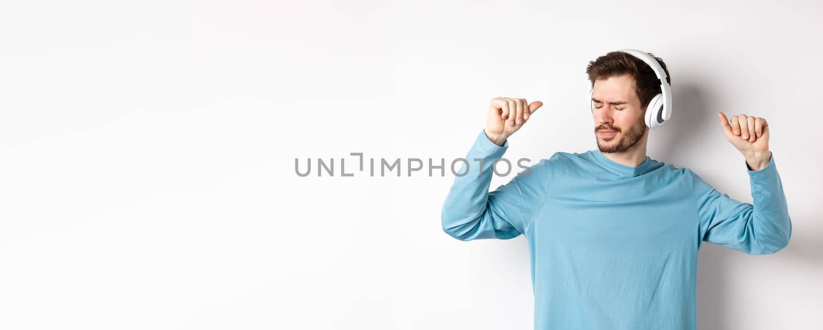 Happy young man having fun in headphones, dancing while listening music in wireless earphones, white background.