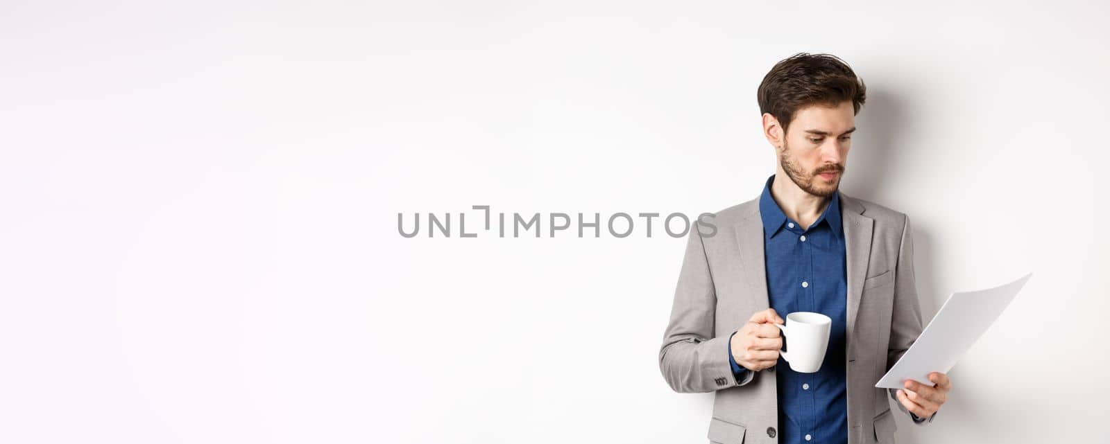 Boss reading documents and drinking morning coffee from mug, looking at paper serious, standing in suit on white background.