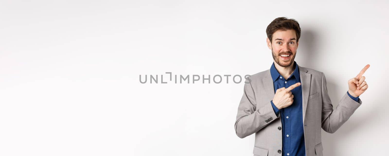 Happy businessman in suit pointing fingers right and smiling excited, showing special deal, standing on white background.