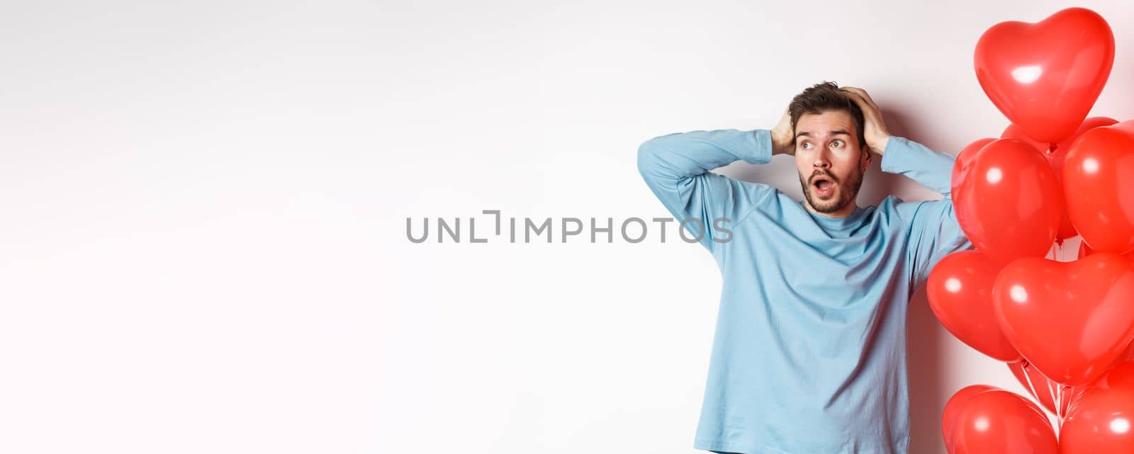 Boyfriend grab head in hands and panic over Valentines day gifts, looking sideways with alarmed face, standing near hearts balloons and thinking of present for lover, white background.