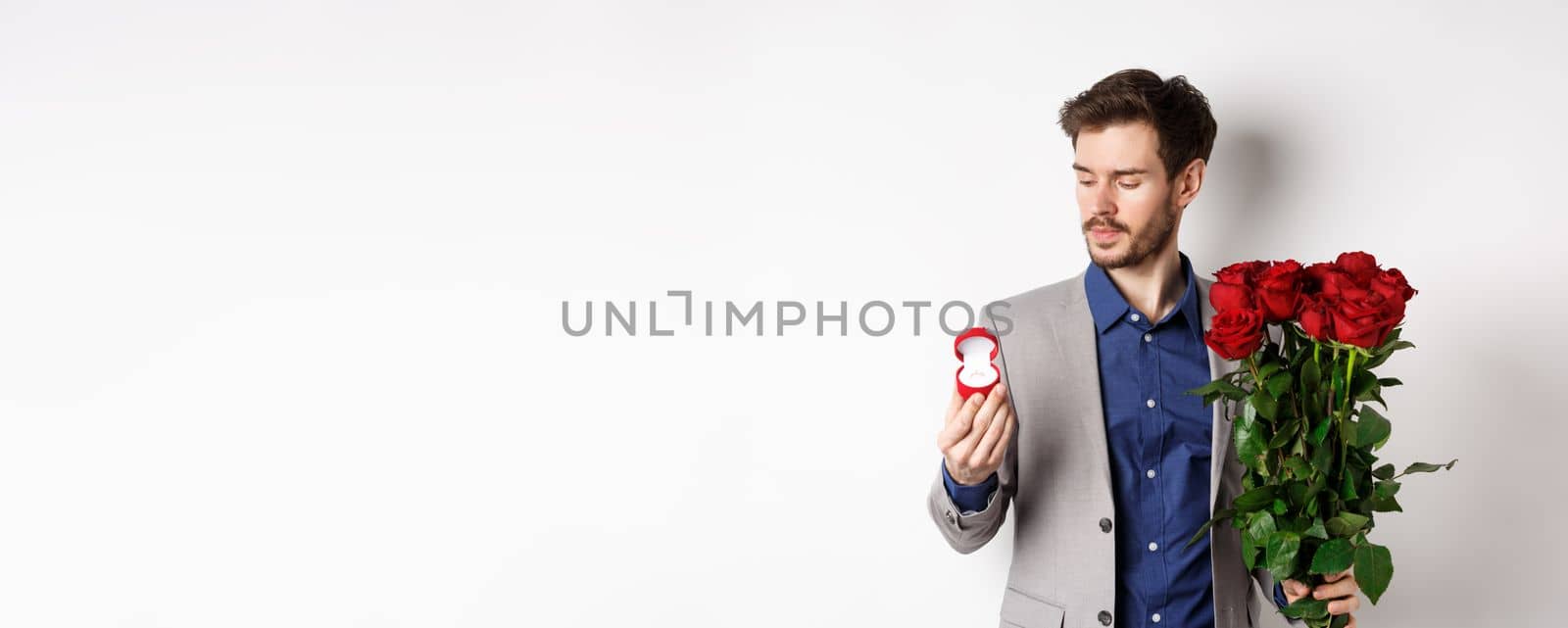 Handsome bearded man in suit looking at engagement ring, making surprise on lovers day, standing with red roses over white background by Benzoix