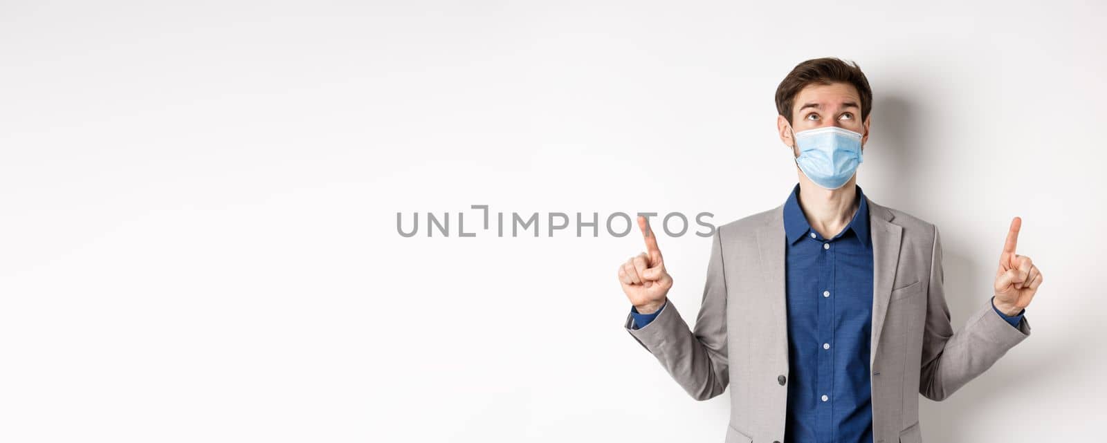 Covid-19, pandemic and business concept. Handsome businessman in medical mask and suit, looking and pointing up with dreamy face, white background by Benzoix