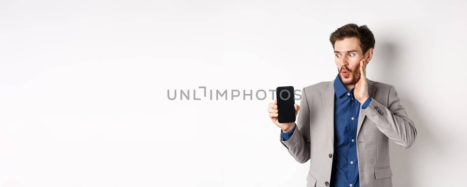 E-commerce and online shopping concept. Amazed male manager in suit looking at mobile phone screen with excited face, standing on white background.