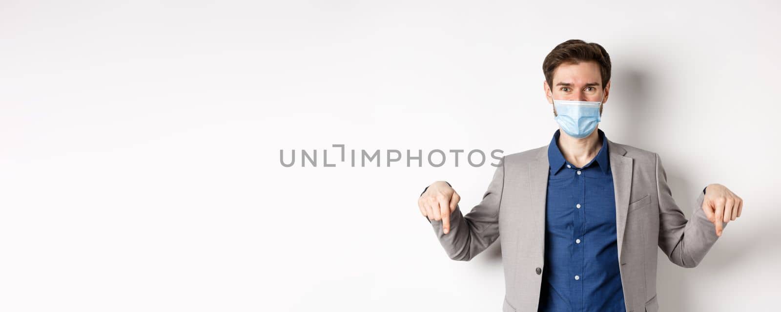 Covid-19, pandemic and business concept. Handsome businessman in medical mask and suit, pointing fingers down and showing company logo, white background by Benzoix