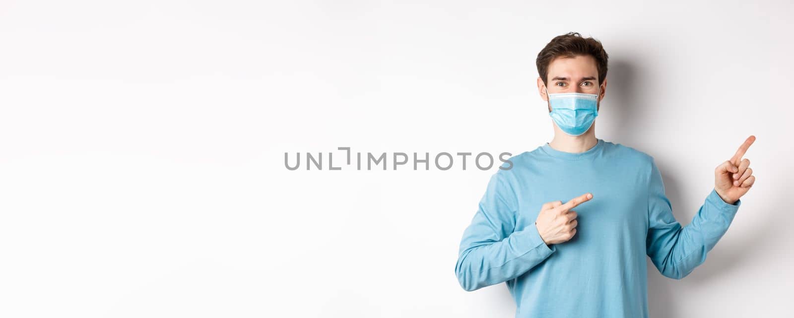 Coronavirus, health and quarantine concept. Handsome caucasian man in face mask pointing fingers right, showing advertisement, standing over white background.