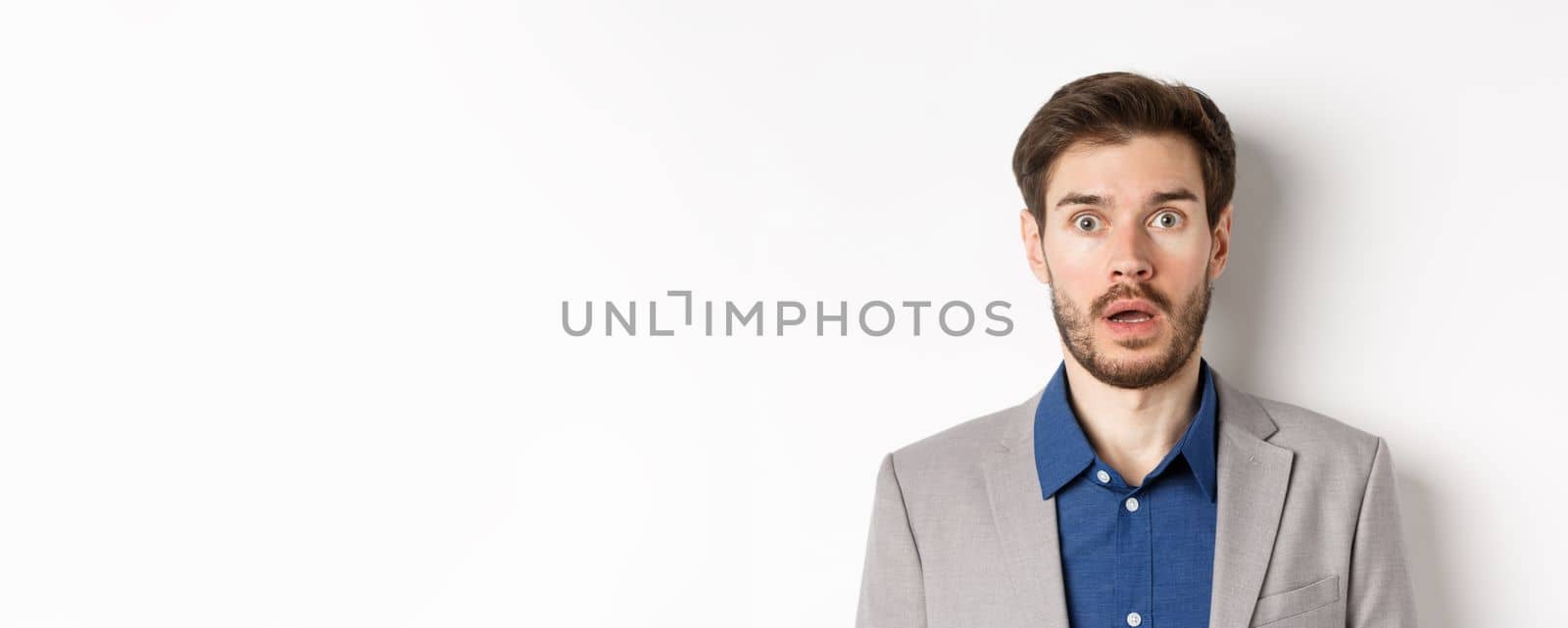 Close up portrait of shocked caucasian man in suit drop jaw, looking speechless at camera, white background.