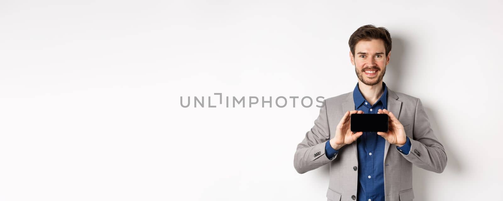 E-commerce and online shopping concept. Happy guy in business suit showing empty smartphone screen horizontally, smiling pleased at camera, white background.