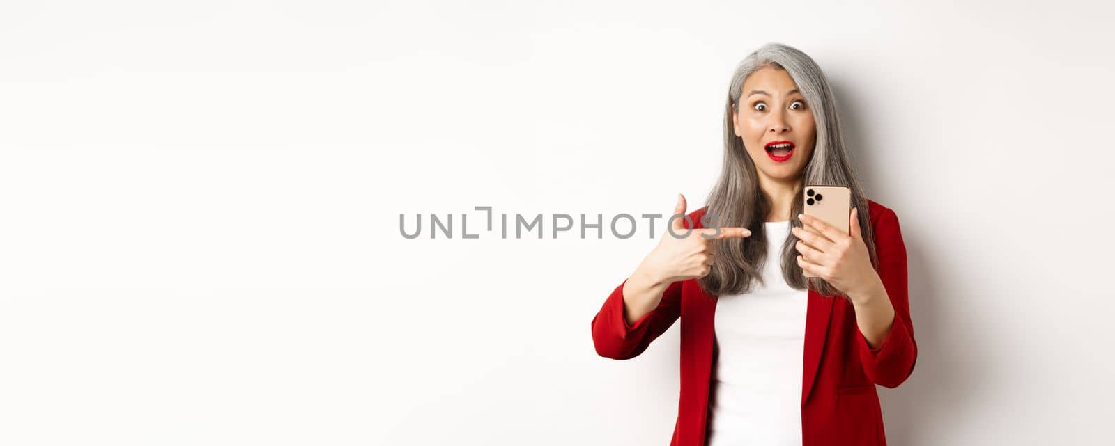 Amazed asian lady in red blazer pointing finger at smartphone, staring impressed at camera, showing something online, white background.