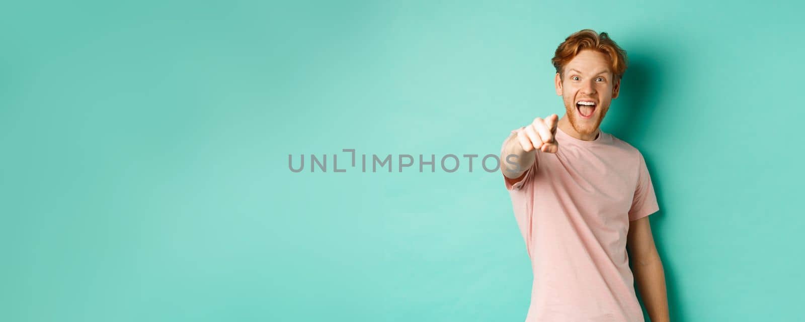 Excited young man with red hair checking out something cool, pointing finger at camera and smiling fascinated, standing over turquoise background by Benzoix