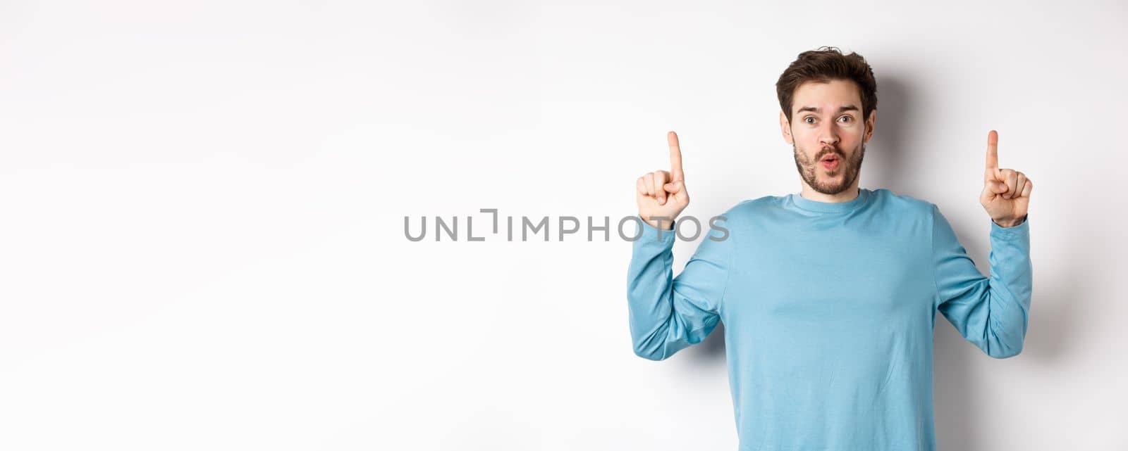 Surprised young man showing top advertisement, pointing up and saying wow at camera, standing over white background.