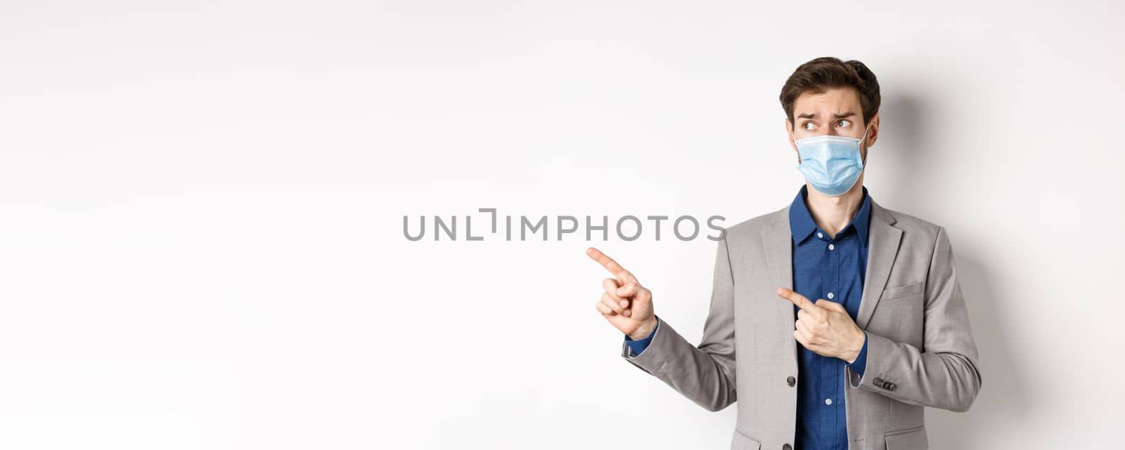 Covid-19, pandemic and business concept. Hesitant businessman in medical mask and suit looking, pointing left with doubtful face, standing on white background by Benzoix