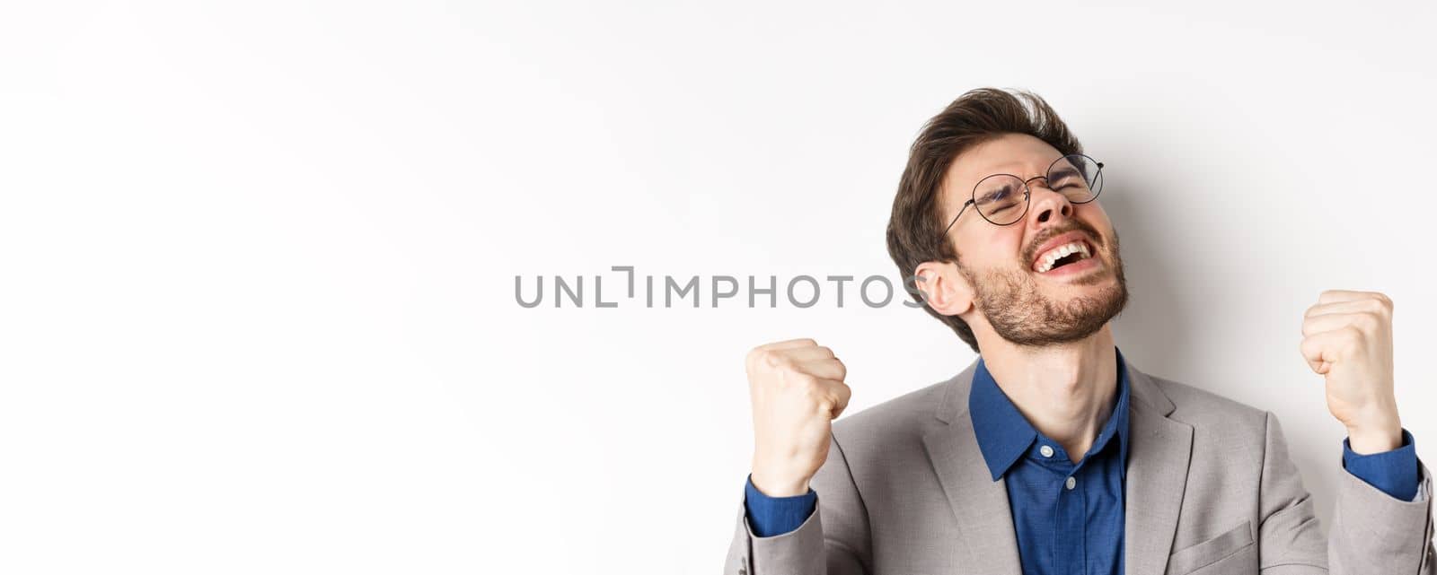 Excited businessman in glasses and suit shouting yes with pleasure and relieved face, shaking fists up, triumphing, winning bet, standing on white background by Benzoix