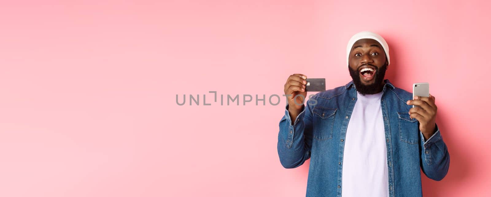 Online shopping. Happy bearded african-american man smiling, showing credit card and making purchase on smartphone, standing over pink background.