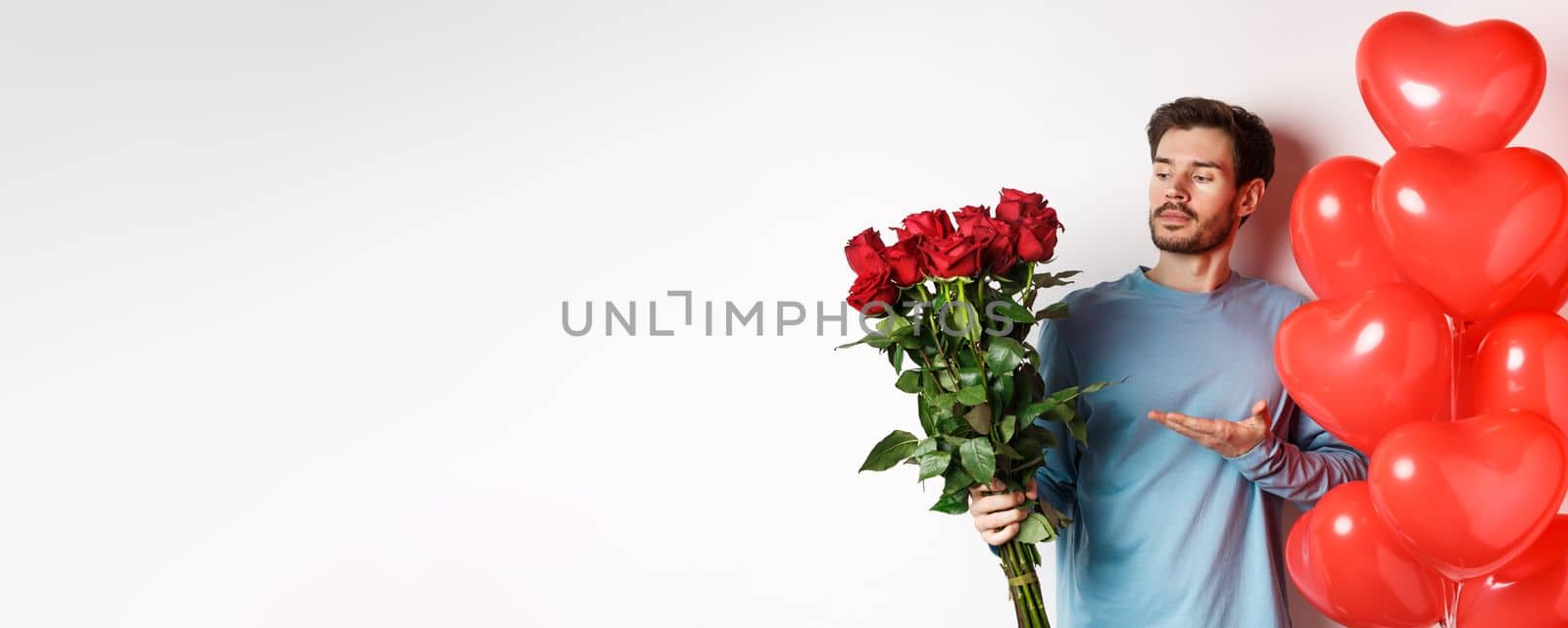 Romantic guy showing bouquet of red roses, pointing at flowers and standing near hearts balloons on Valentines day, prepare gifts for his lover, white background.