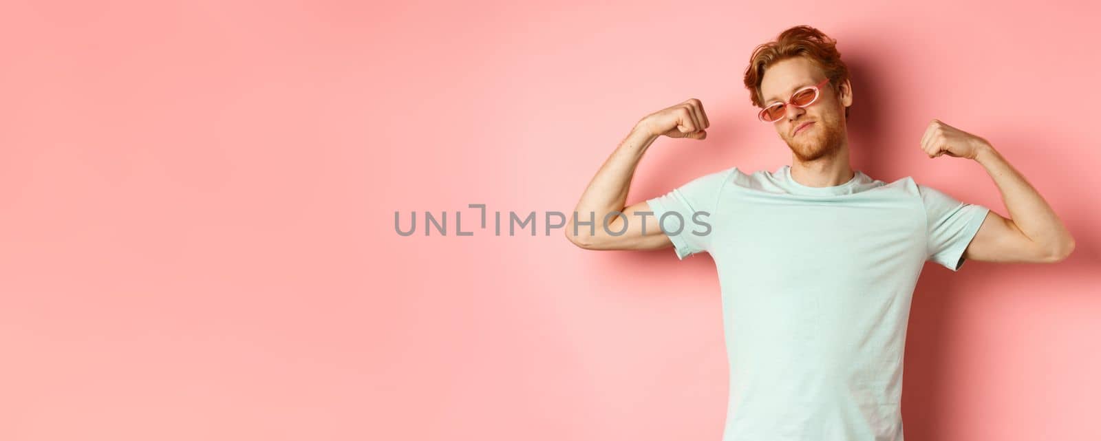 Confident young man with red hair, wearing summer sunglasses and t-shirt, showing strong and fit body muscles, flex biceps and staring cool at camera, pink background. Workout and gym concept