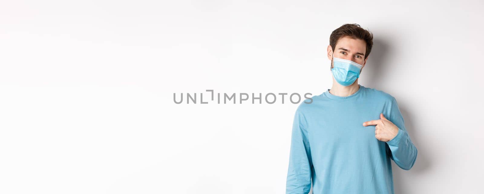 Covid-19, health and quarantine concept. Smiling caucasian man in medical mask pointing at himself, volunteering, standing over white background.