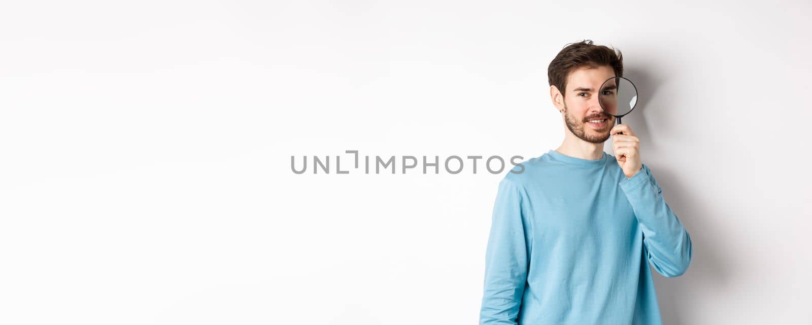 Smiling young man detective looking through magnifying glass, searching for promo offer, standing on white background.