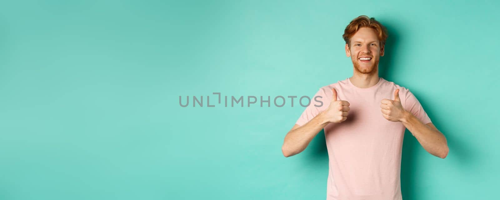 Cheerful bearded man with red hair showing thumbs-up, like and approve something, praising promo, standing against turquoise background.