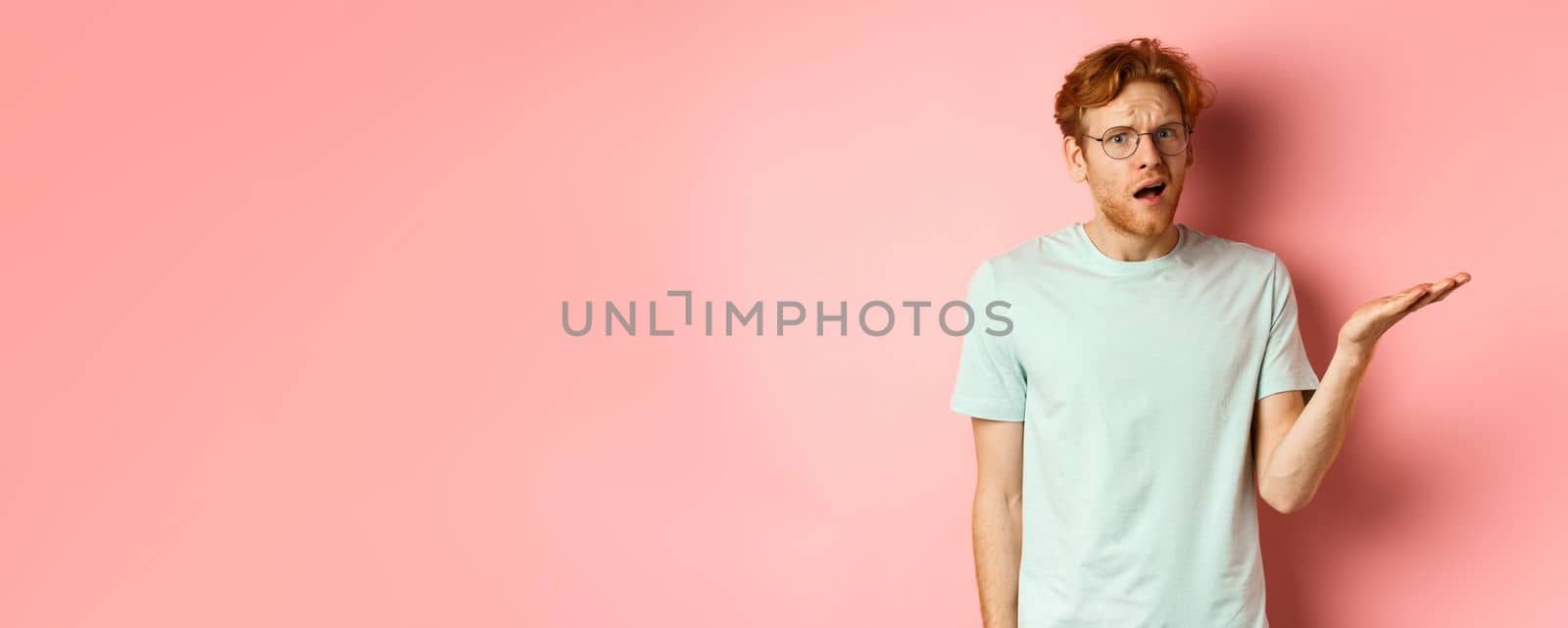 Portrait of handsome redhead man in glasses looking confused, open mouth and stare at camera, cant understand nothing, standing over pink background.