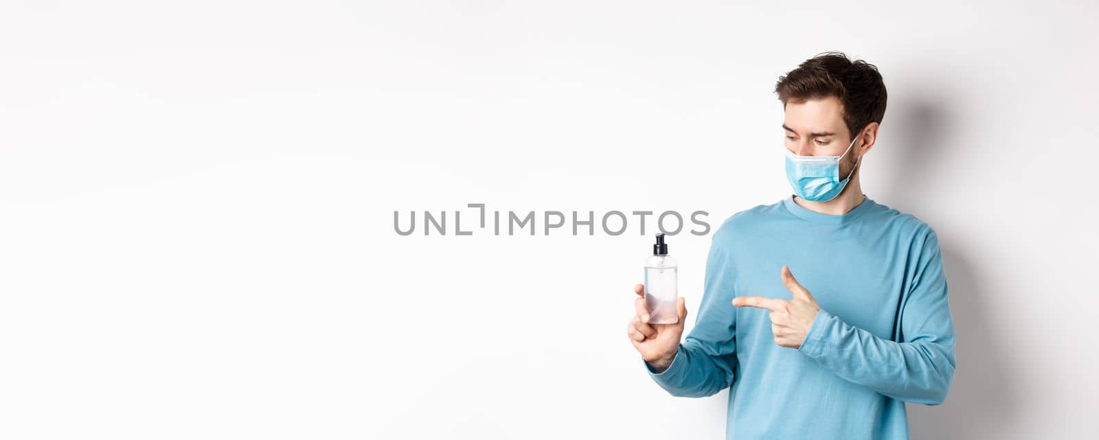 Covid-19, health and quarantine concept. Young man pointing and looking at hand sanitizer bottle, showing good antiseptic, white background by Benzoix