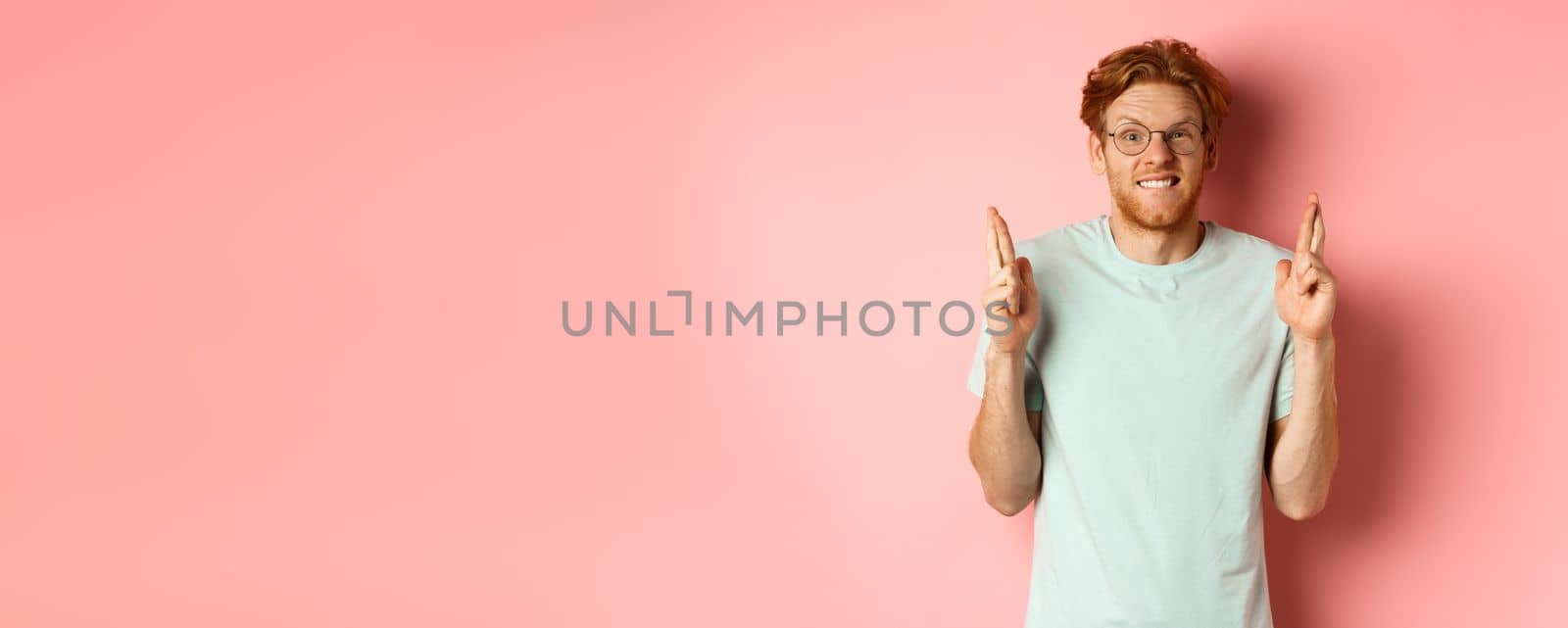 Worried redhead man waiting for results, expecting something with fingers crossed, biting finger and looking at something risky, standing over pink background by Benzoix